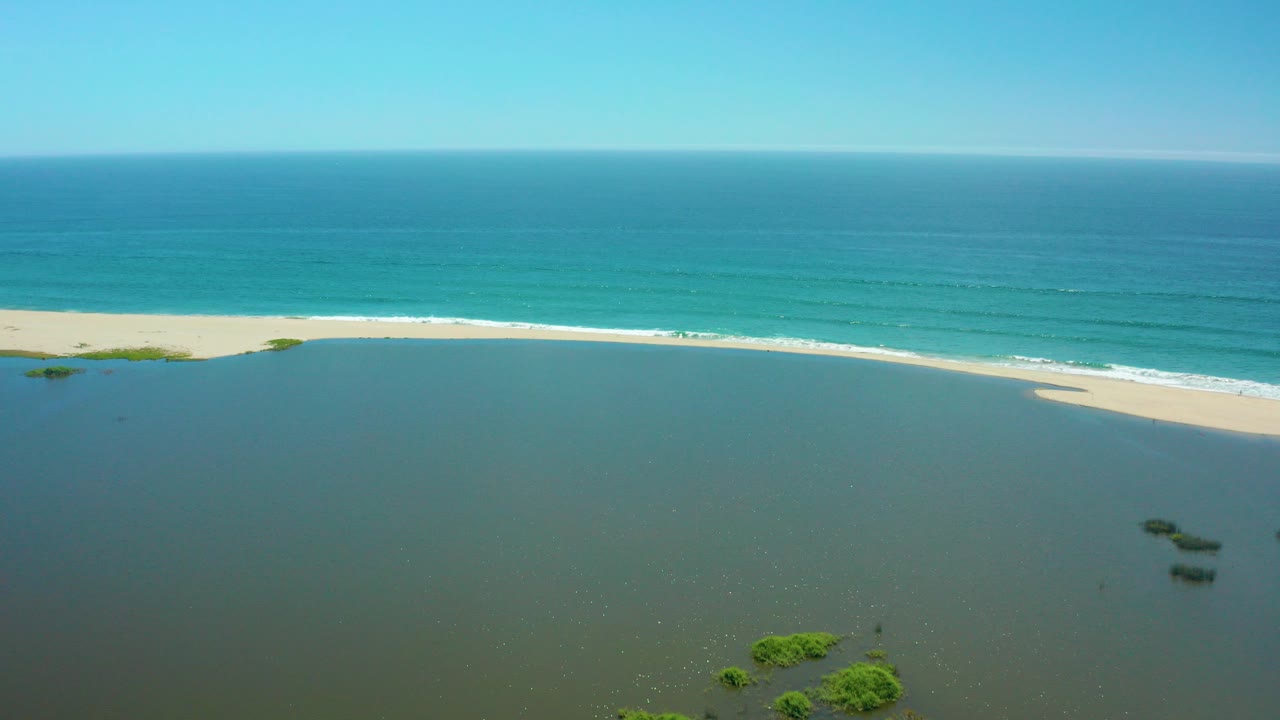 Aerial frame from San Jose Del Cabo showing shallow lagoon water in front, a pale sandbar, turquoise ocean beyond, scattered green mangroves, and no visible people.