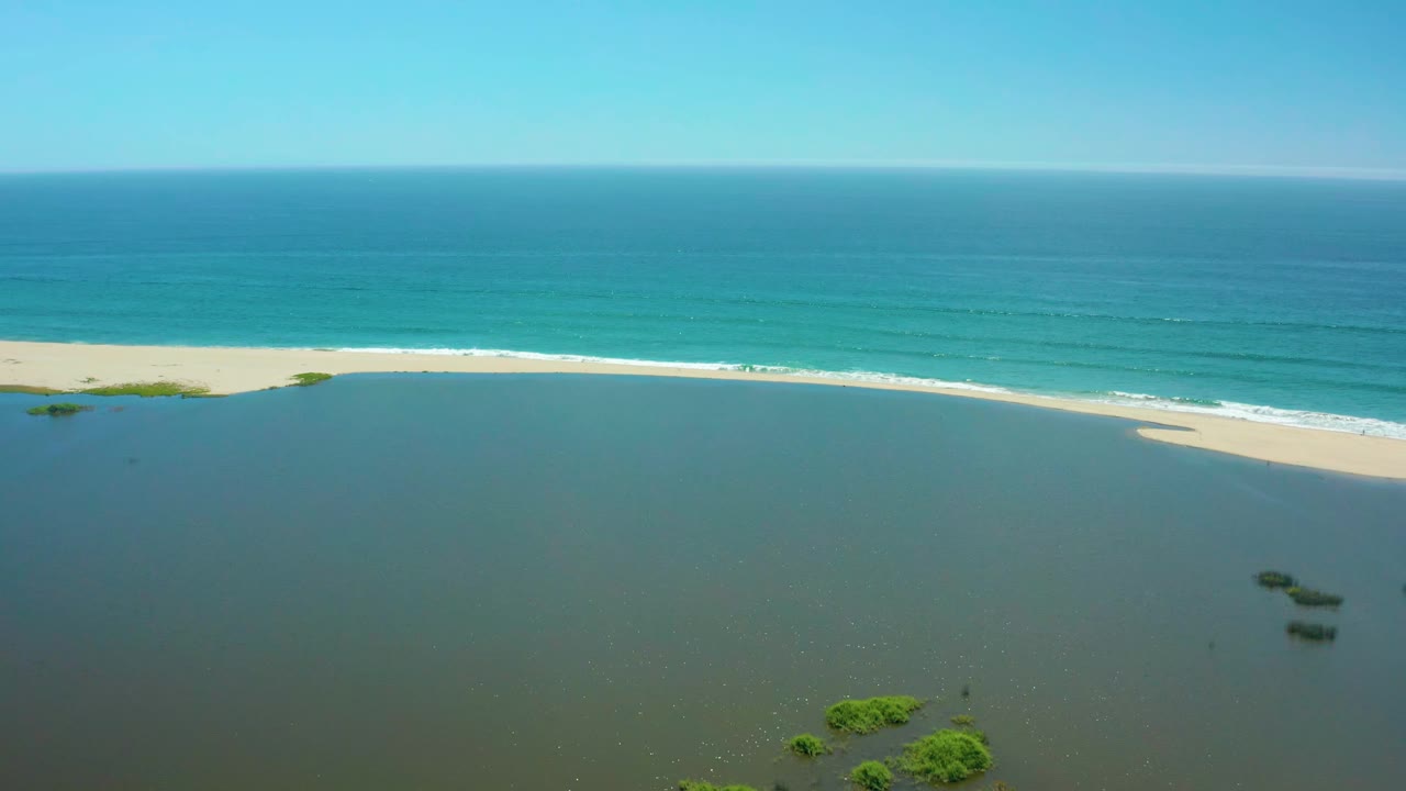 A second aerial frame of San Jose Del Cabo shoreline shows the lagoon filling the lower frame, the sandbar crossing the middle, ocean bands behind it, and no visible people.