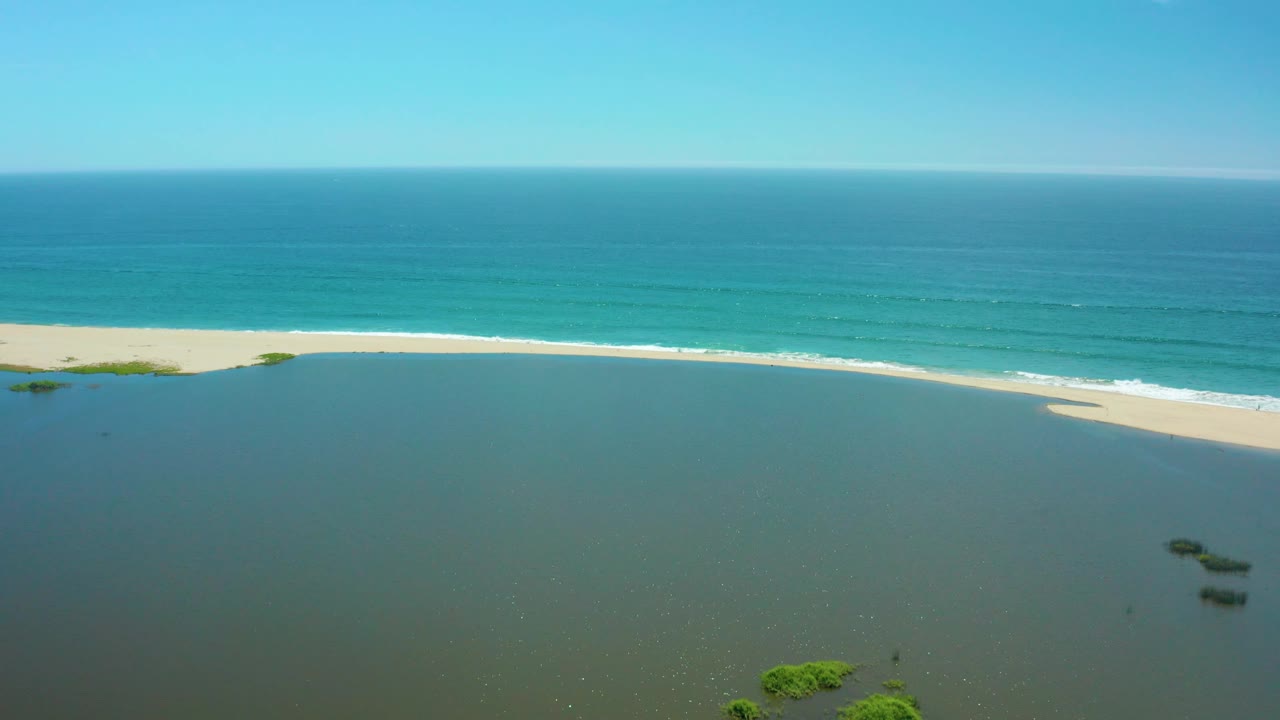 Aerial view over San Jose Del Cabo lagoon water with a thin pale sandbar, turquoise ocean, small surf streaks, foreground mangrove clumps, and no visible people.