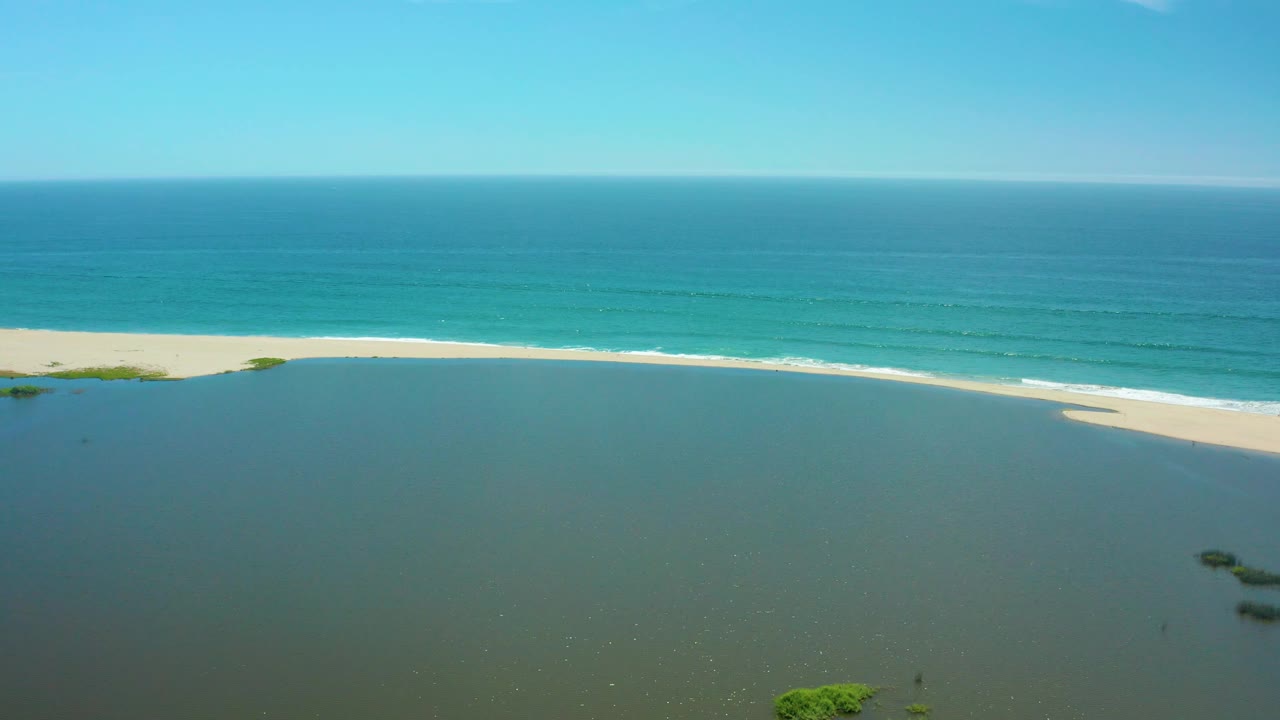 San Jose Del Cabo aerial shoreline frame with shallow lagoon water in the foreground, a bright sand edge, layered blue ocean in the background, and no visible people.