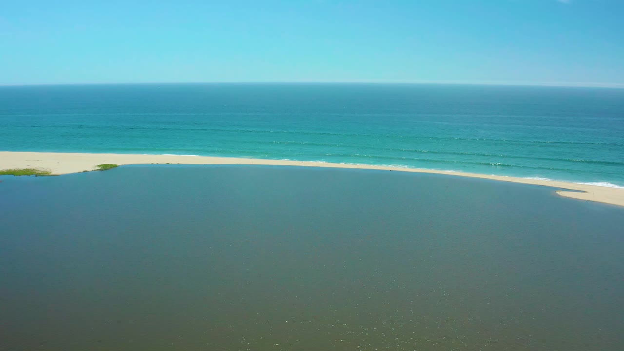 Wide aerial frame in San Jose del Cabo with still lagoon water across the foreground, a bright sandbar through the middle, ocean surf beyond it, and no visible people.