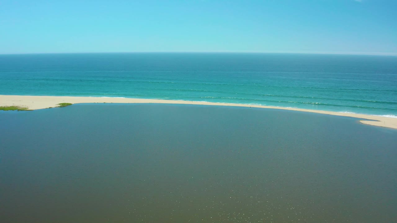 Aerial coastal frame in San Jose del Cabo showing muted lagoon water below, a thin pale sandbar with a small curved shoal, turquoise ocean, and no visible people.