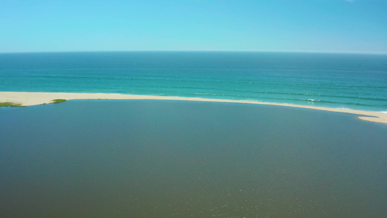 San Jose del Cabo aerial view with broad green lagoon water, a horizontal strip of pale sand, turquoise ocean bands, clear blue sky, and no visible people.