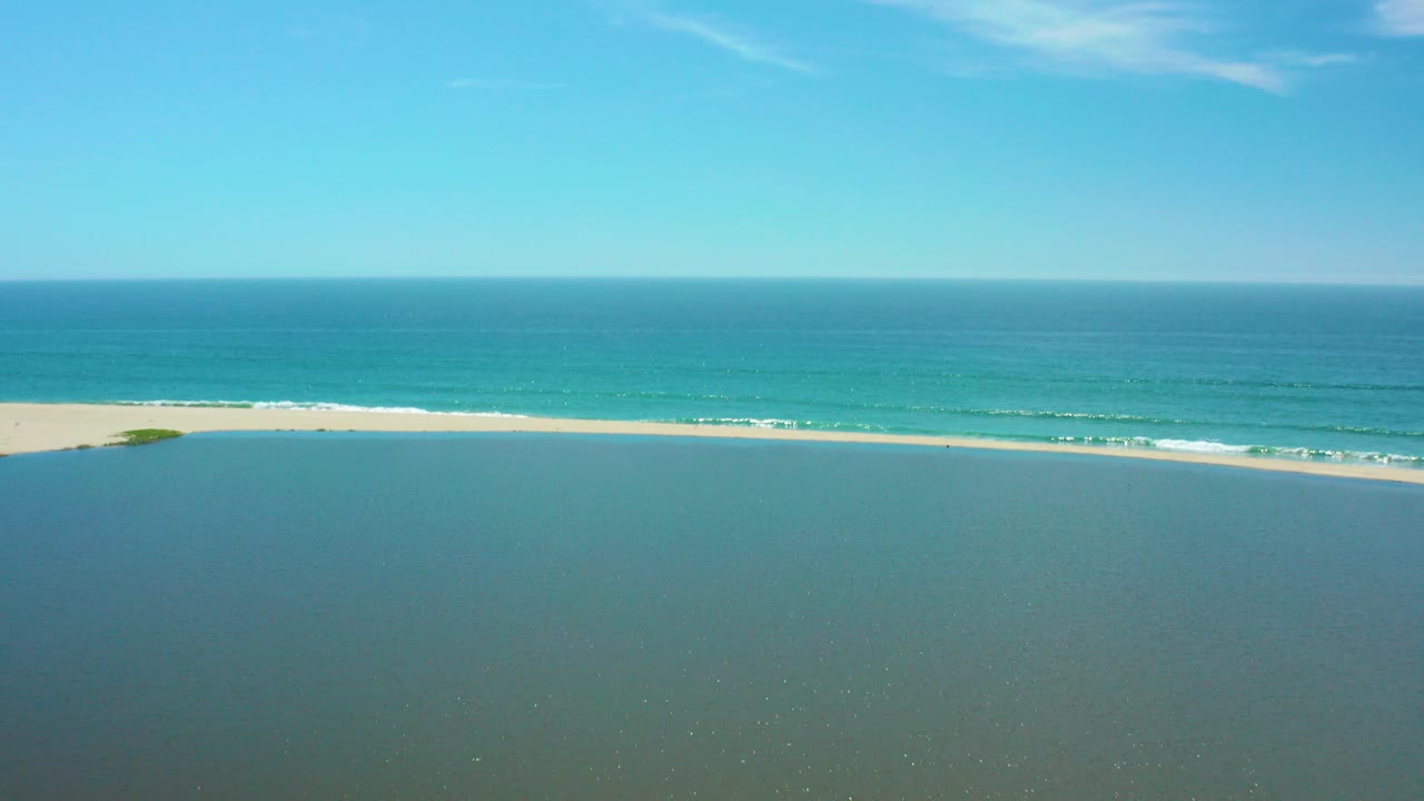 Wide aerial view of lagoon, sandy shore, ocean waves, and blue sky.