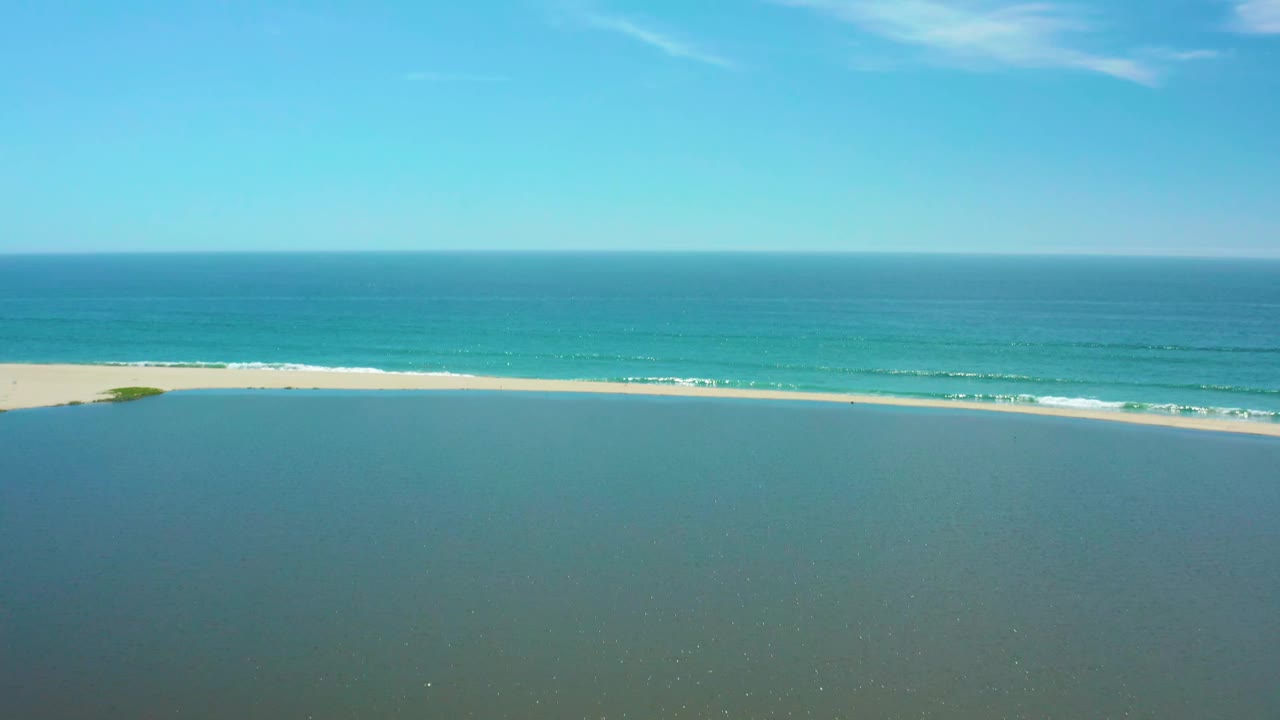 San Jose del Cabo aerial view with more open lagoon foreground, a pale sand strip, thin white surf lines, turquoise sea, clear sky, and no visible people.