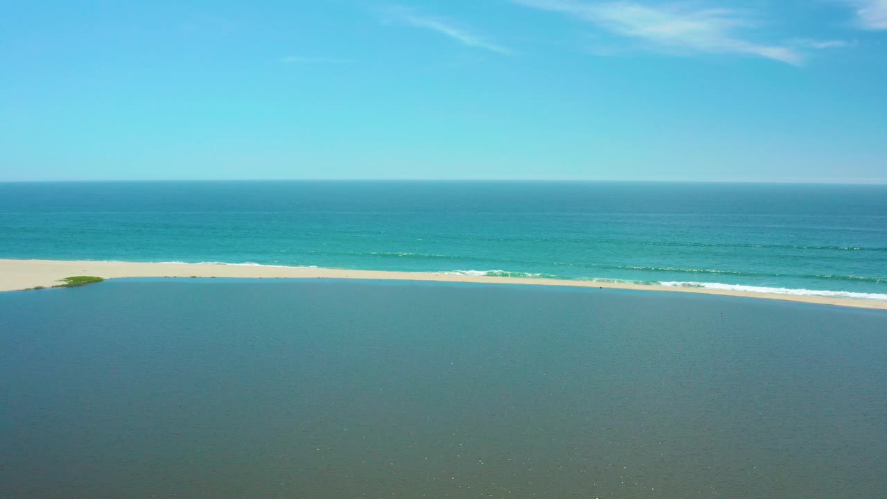 San Jose del Cabo coastal aerial with calm lagoon filling the lower frame, a narrow sandbar, darker turquoise ocean beyond, light surf foam, and no visible people.