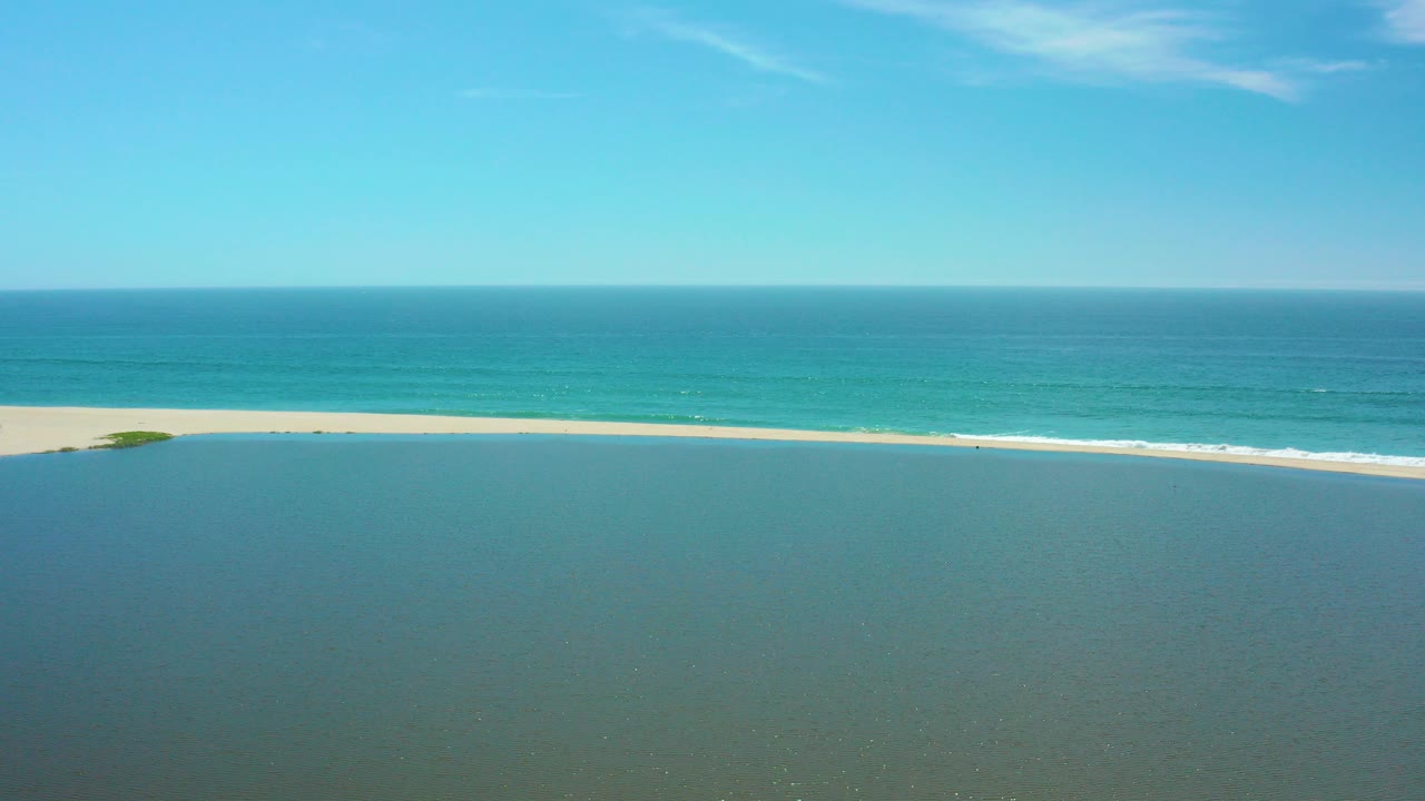 Aerial frame over San Jose del Cabo with greenish lagoon water in the foreground, a pale sand beach, blue ocean beyond it, clear sky, and no visible people.
