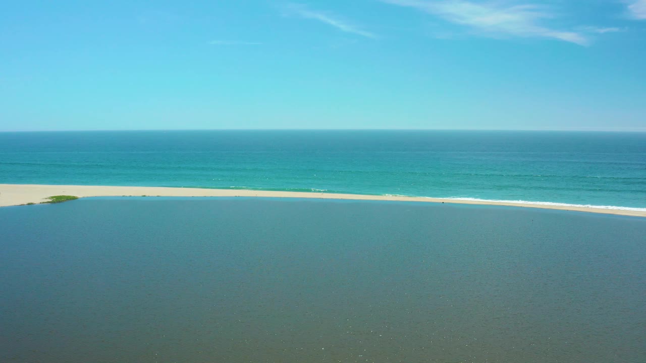 San Jose del Cabo aerial frame with a long pale beach stretching across the middle, darker lagoon water below, turquoise ocean above, and no visible people.