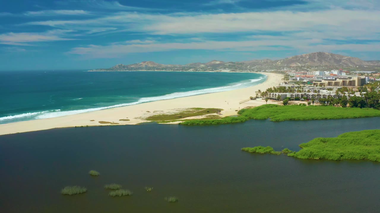 Early runtime frame of San Jose Del Cabo showing mangrove patches scattered across dark lagoon water, with a bright sand beach, curved surf line, distant buildings, hills, and no visible people.