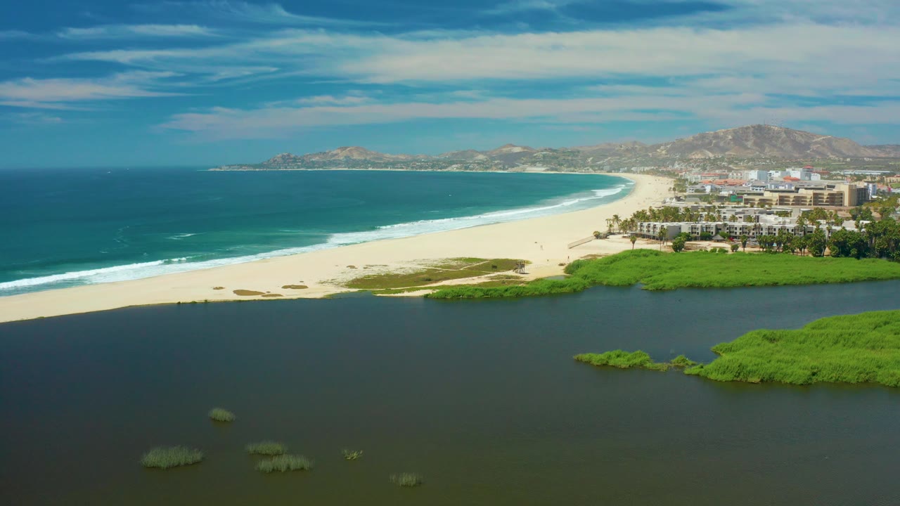 Mid-clip aerial frame in San Jose Del Cabo with dark water and small mangrove clumps in the foreground, a broad empty sand beach, blue ocean, shoreline buildings, hills, and no visible people.