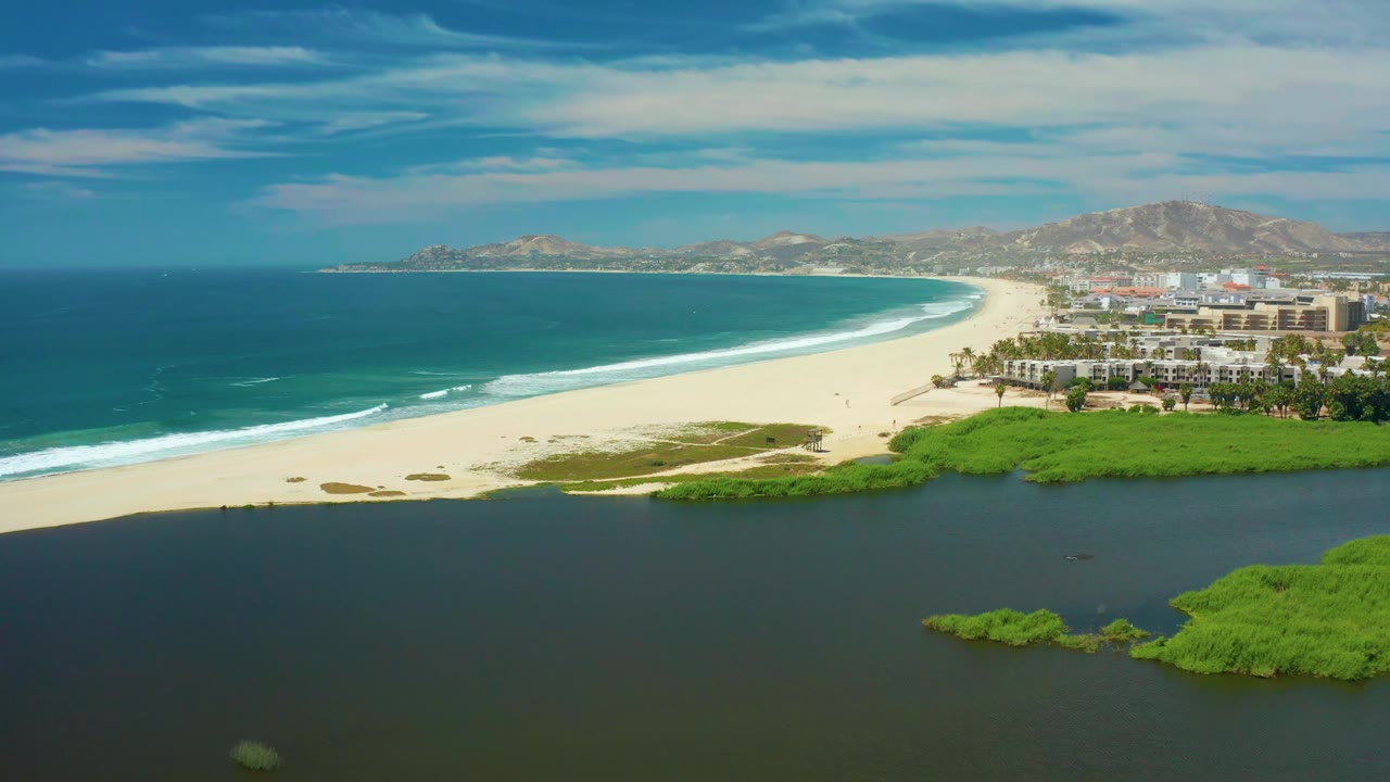 Wide San Jose del Cabo aerial with the beach curving between the dark lagoon and blue ocean, low white surf on the shore, green vegetation patches, and no visible people.