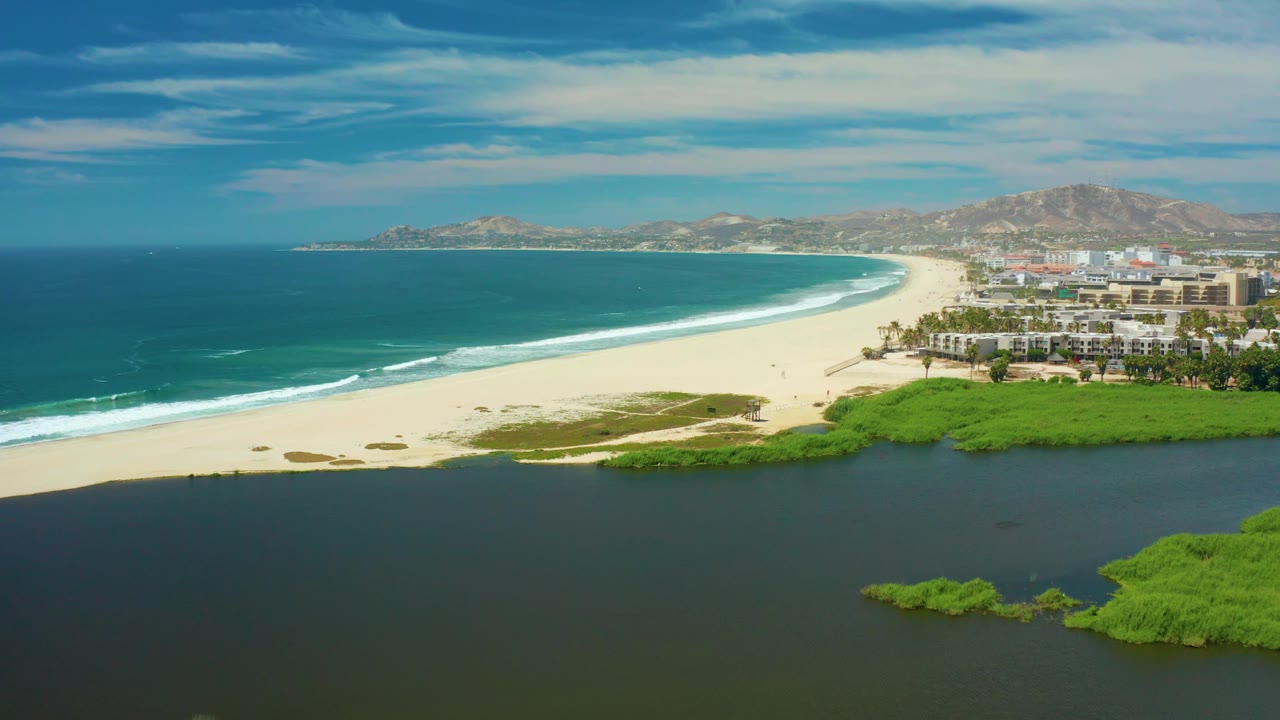 San Jose del Cabo coastal aerial showing blue ocean at left, a bright strip of beach, a dark lagoon below, green shoreline vegetation at right, distant hills, and no visible people.