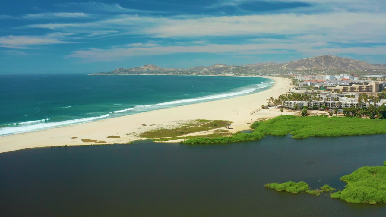 Aerial frame over San Jose del Cabo with ocean waves reaching the sandy beach, lagoon water across the lower frame, green vegetation near shore, distant hills, and no visible people.