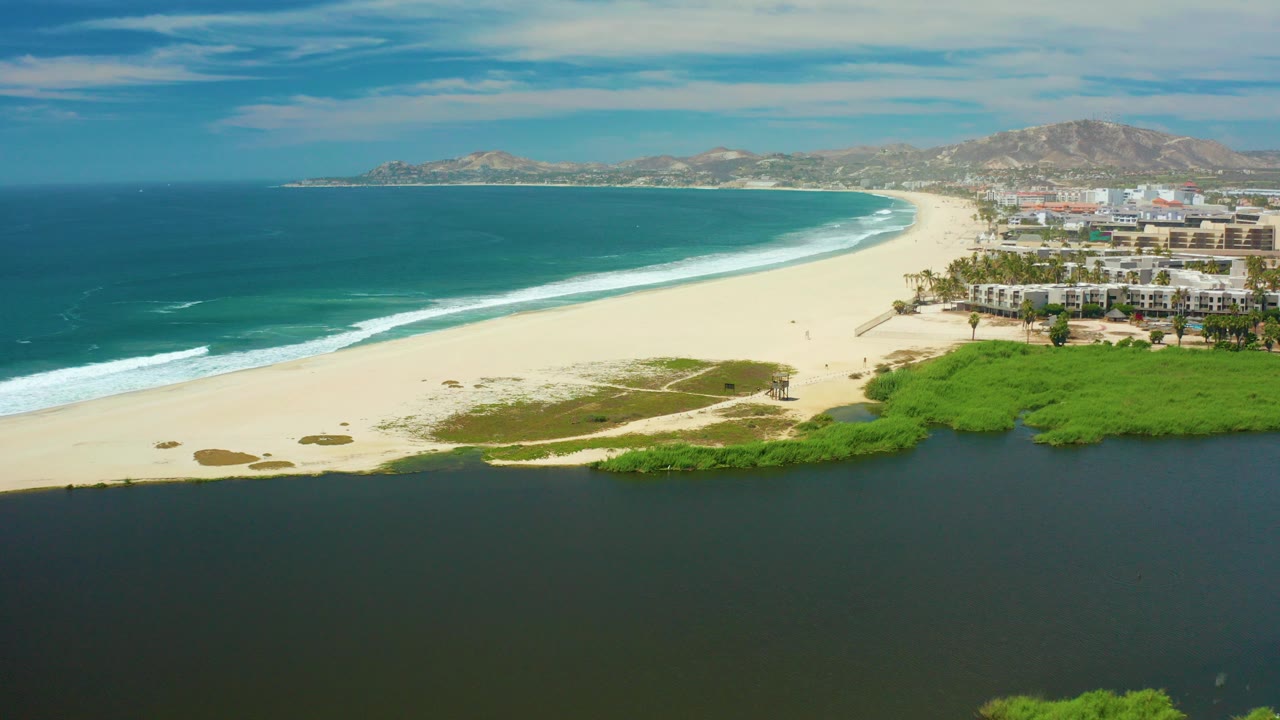 San Jose del Cabo aerial frame with dark lagoon water in the foreground, pale beach and green vegetation in the middle, ocean surf curving left to right, beachfront buildings on the right, distant hills, and no visible people.
