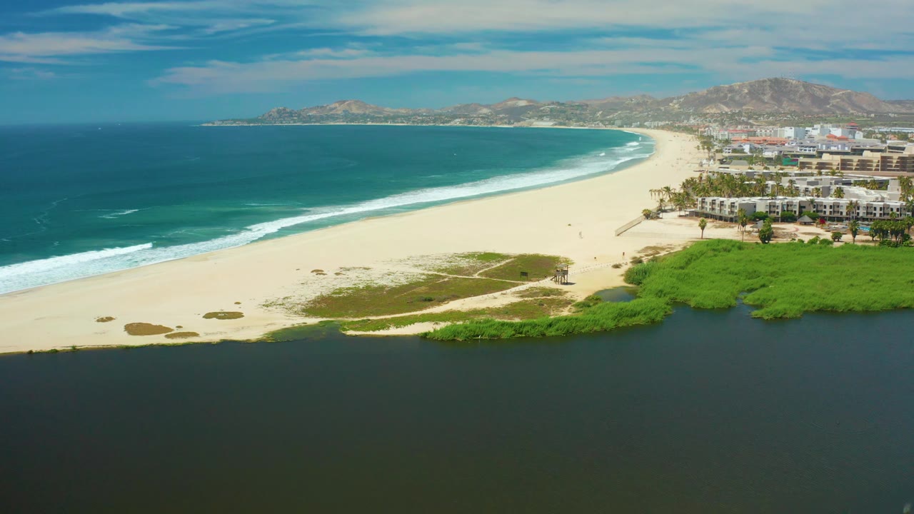 San Jose del Cabo beach aerial with turquoise water on the left, white surf tracing the sand, lagoon and greenery along the lower edge, beachfront buildings on the right, hazy hills beyond, and no visible people.