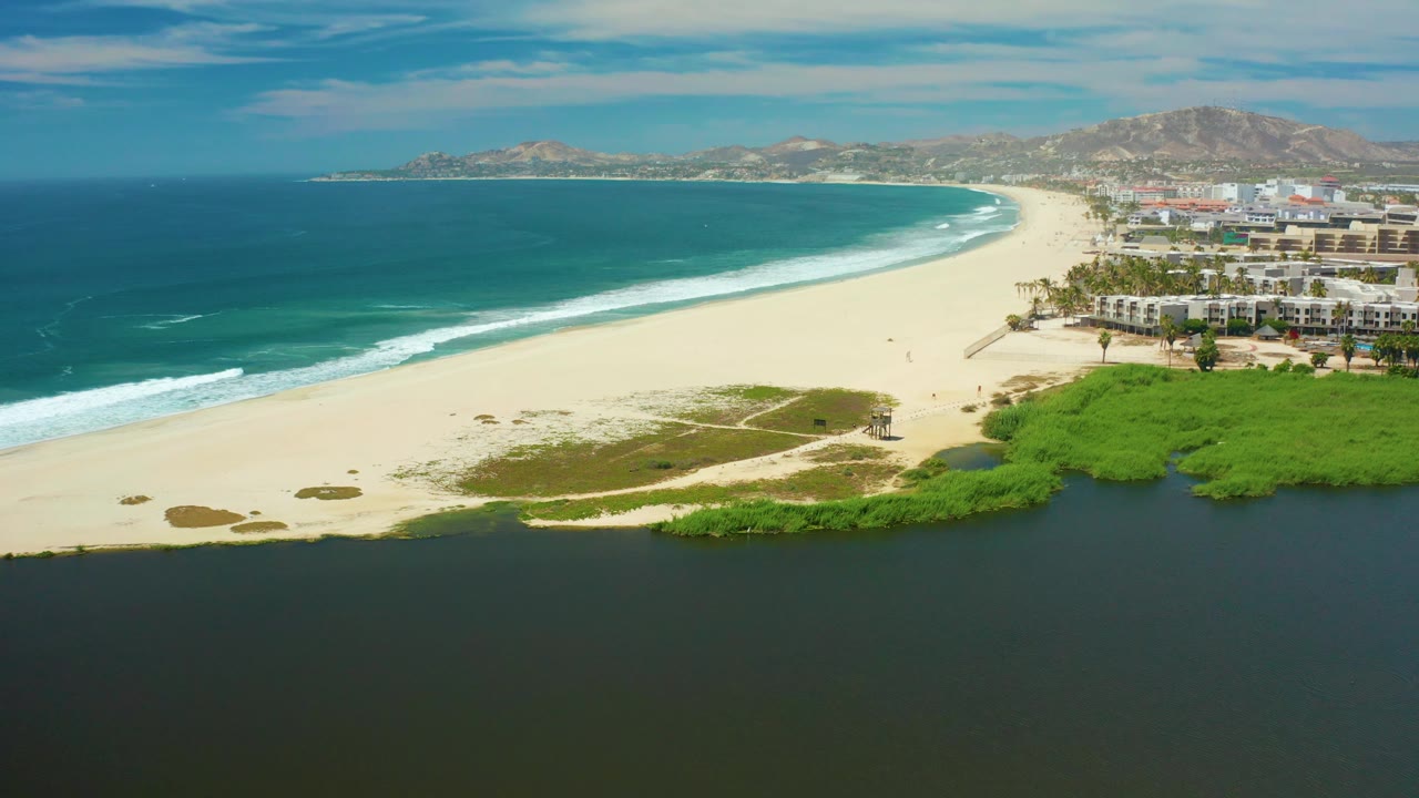 Coastal aerial with dark lagoon water below, pale sand, surf, buildings, and mountains.