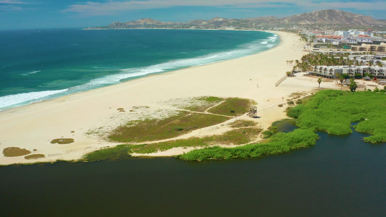 Wide aerial frame over San Jose Del Cabo showing a dark lagoon below, pale beach, blue ocean surf, coastal greenery, distant buildings, hills, and no visible people.