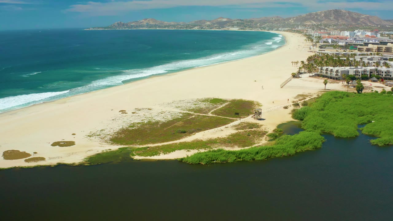 Aerial coastline with sandy beach, dark lagoon water, ocean waves, and low greenery.