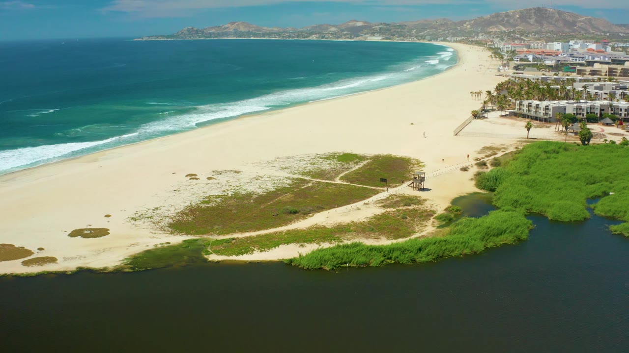 San Jose Del Cabo aerial frame showing a sweeping beach curve, turquoise ocean, white waves, dark lagoon edge, green dune vegetation, beachfront buildings, and no visible people.