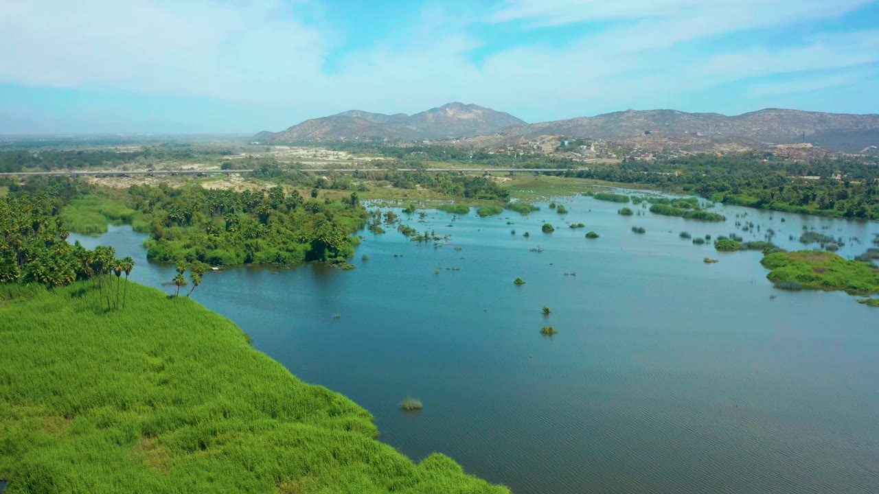 Aerial frame in San Jose del Cabo showing more open lagoon water in the foreground, green vegetation islands through the center, a low mountain ridge on the horizon, and no visible people.