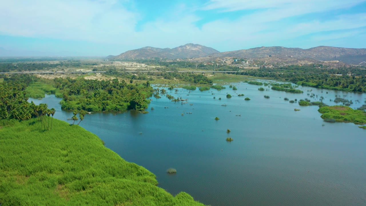 Mid-clip aerial frame over the San Jose del Cabo lagoon with a vivid green slope in the lower left, darker tree clusters along the water, scattered islets, distant mountains, and no visible people.