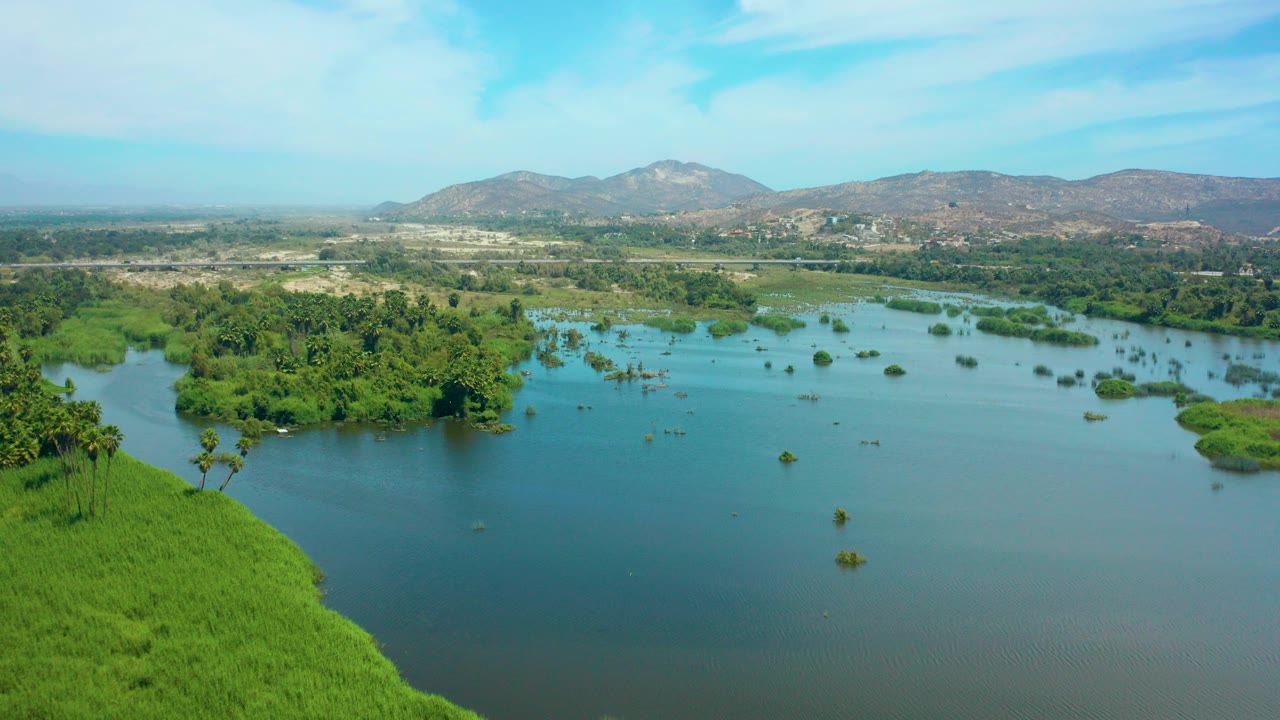 Aerial frame from San Jose del Cabo showing calm blue-green water, small tree-covered islands, a bright green bank in the lower left, distant hills, and no visible people.