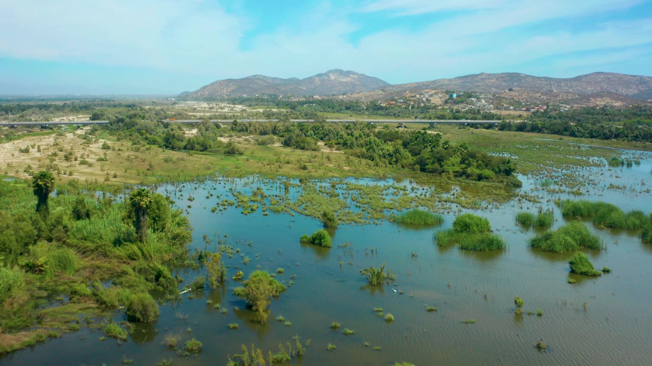 Aerial frame over San Jose del Cabo showing blue-green floodwater, scattered wetland shrubs, a background road, distant hills, and no visible people.