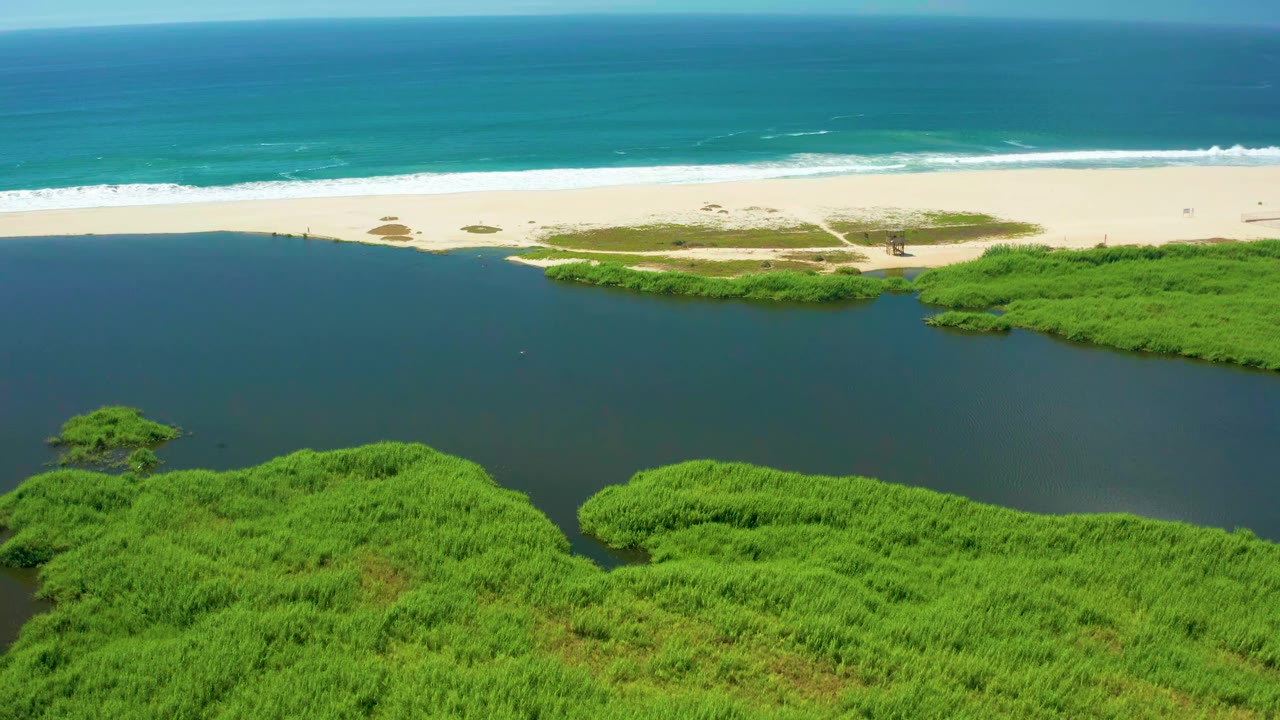 At 0:01, the aerial view in San Jose Del Cabo shows the lagoon slightly more centered, with mangrove edges framing the lower side, white beach sand above it, and no visible people.
