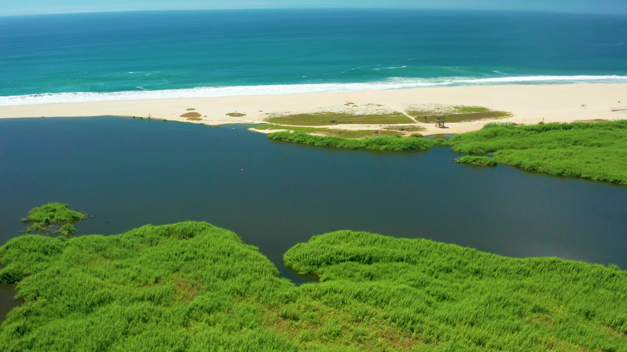 At 0:02, the San Jose Del Cabo aerial angle shows a broad dark lagoon below the pale sandbar, with vivid green mangroves at the lower edge and no visible people.