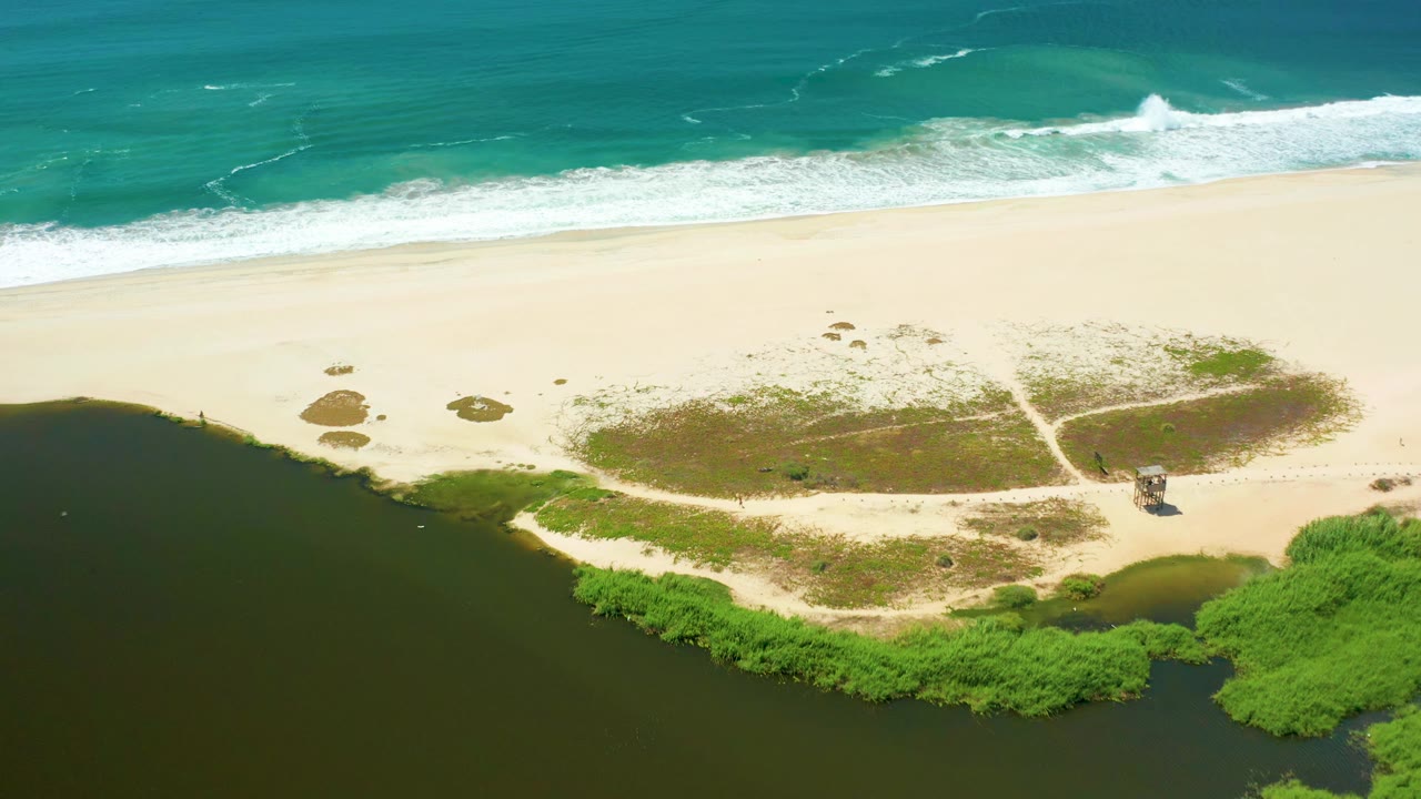 Aerial view in San Jose Del Cabo with white surf bands near the top, pale sand across the center, a dark river estuary at the bottom, sandy paths, and no visible people.
