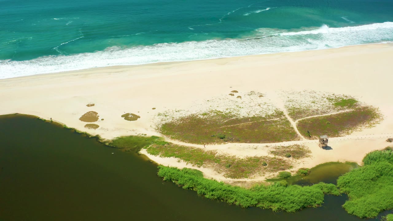 San Jose Del Cabo aerial frame showing a long white wave break, open sand, scattered scrub patches, dark estuary water, green vegetation, and no visible people.