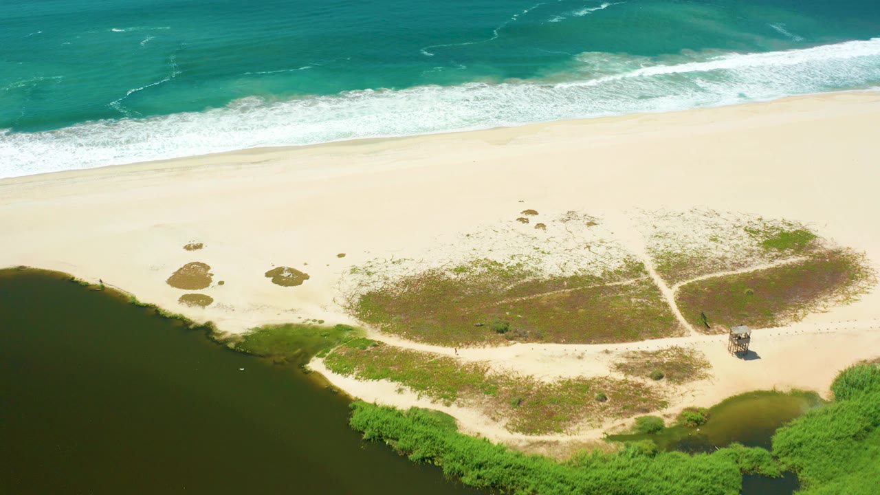 Aerial San Jose Del Cabo shoreline frame with ocean foam along the upper edge, broad empty sand, dune scrub, dark estuary water, green plants, and no visible people.