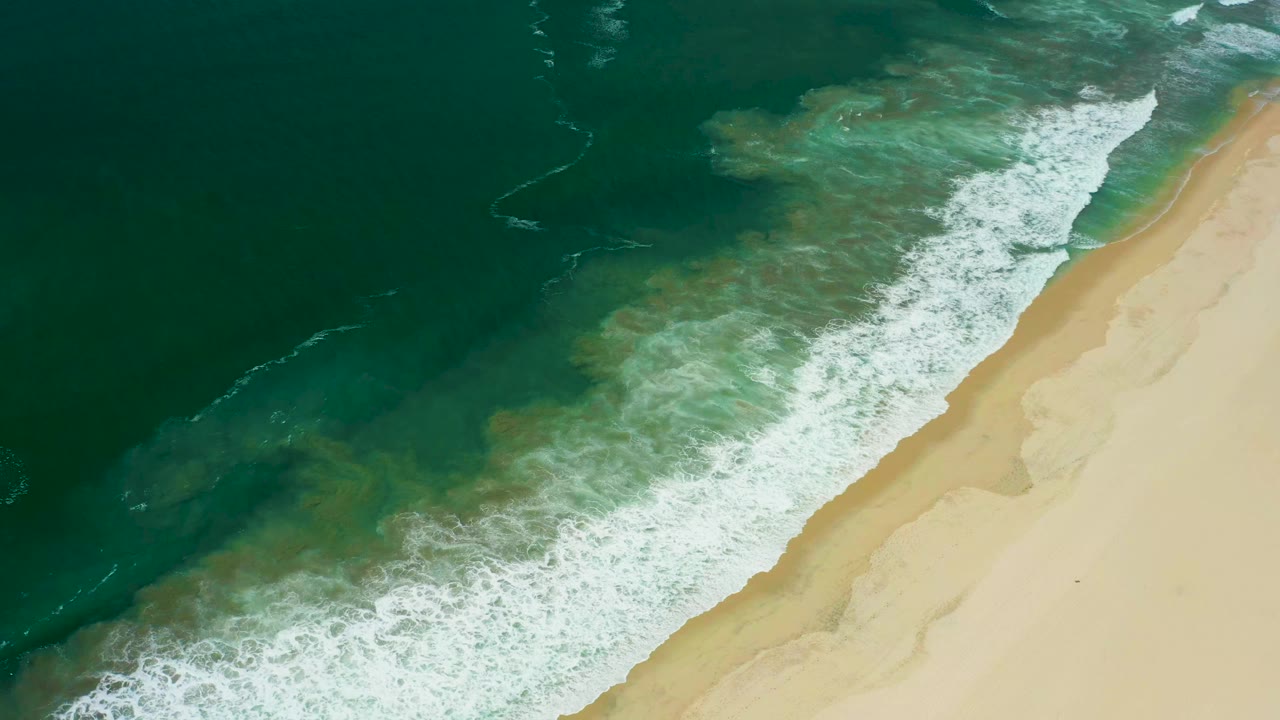 San Jose del Cabo shoreline seen from above with a broad white foam band closer to the sand and darker green water filling the left side, with no visible people.
