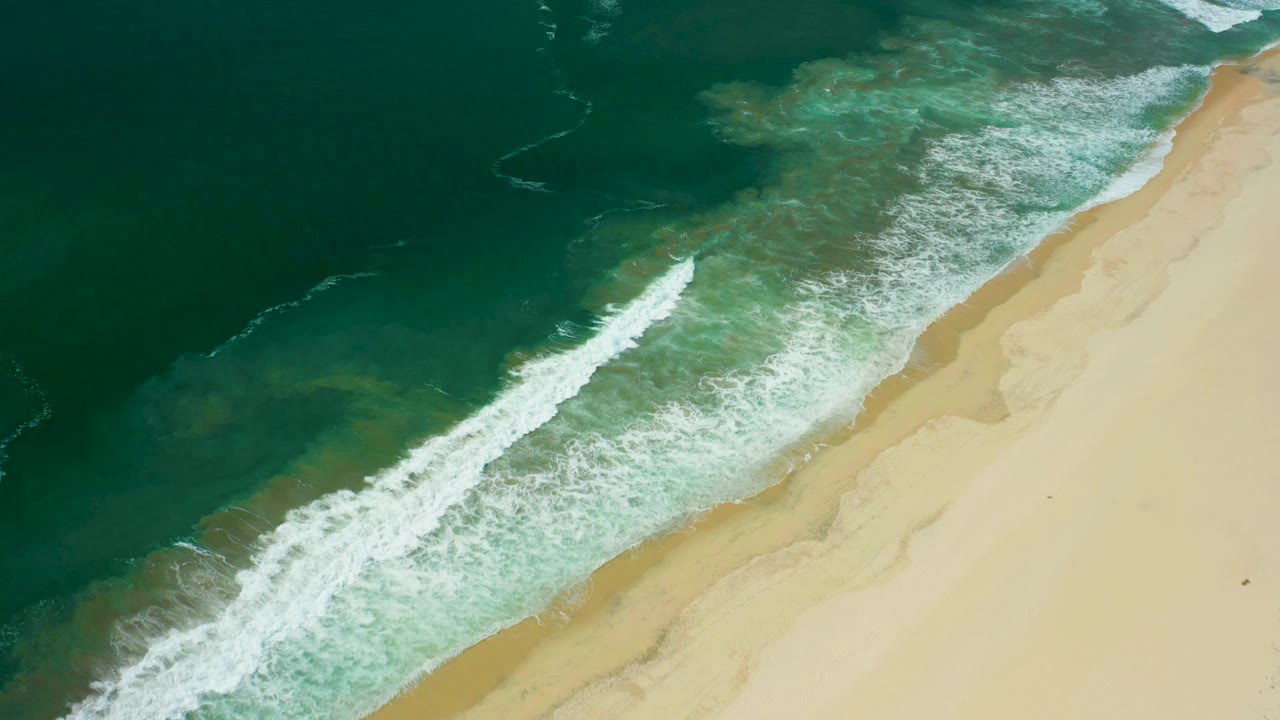 Aerial San Jose del Cabo shoreline view with a longer white breaker pushing toward the pale beach and no visible people.