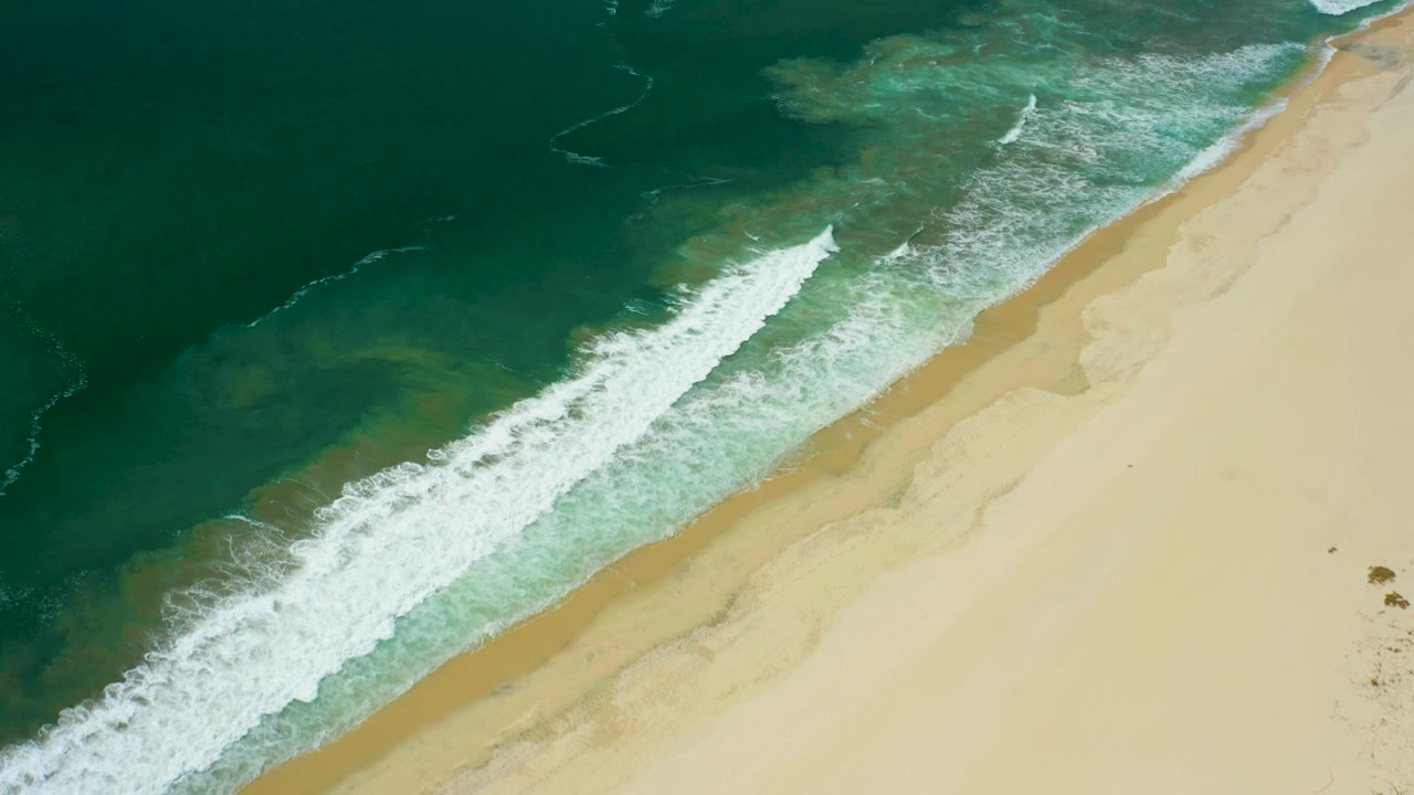 San Jose del Cabo aerial shoreline frame with the ocean occupying most of the left side, a foamy breaker in the center, and empty pale sand to the right.