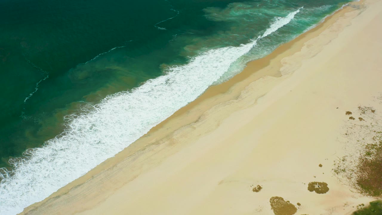 White foam advances farther along the San Jose Del Cabo shoreline, with dark green water offshore and pale sand filling the lower right, no visible people.
