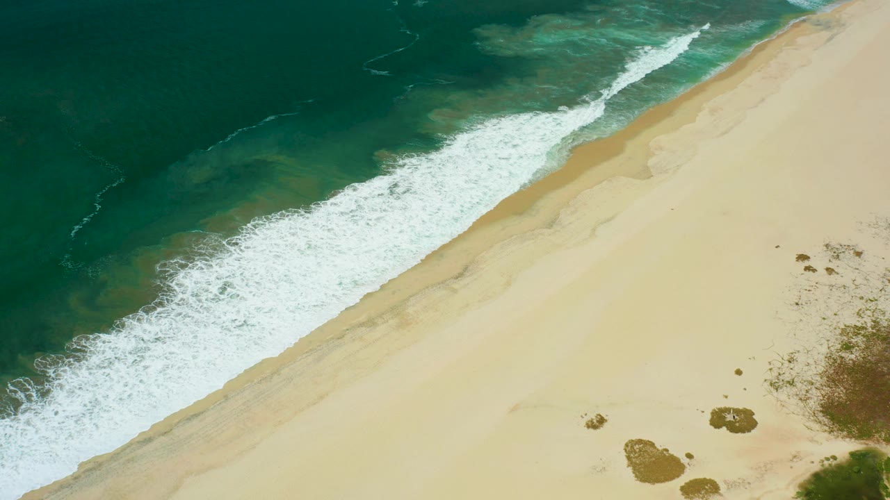 The wave edge stretches across the frame off San Jose Del Cabo, with white foam against tan sand and small darker rock patches near the right side, no visible people.