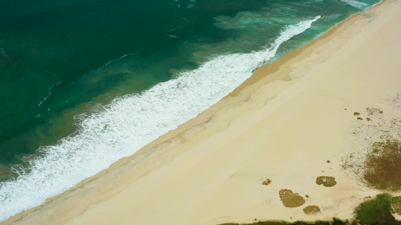A broad band of white surf curls onto the San Jose Del Cabo beach, with darker ocean in the upper left and open sand in the lower right, no visible people.