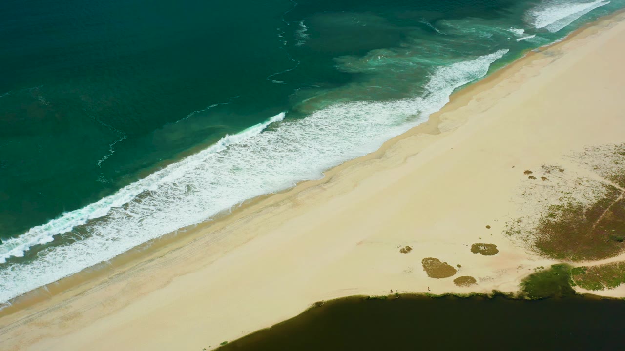 Aerial frame in San Jose del Cabo with a broad band of white surf pushed slightly farther along the sand, dark lagoon water below, and no visible people.