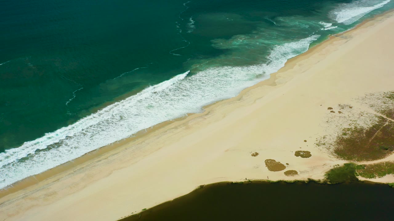 San Jose del Cabo shoreline from above, with teal water filling the upper frame, foam tracing the beach, and small vegetation patches near the dark lagoon, no visible people.