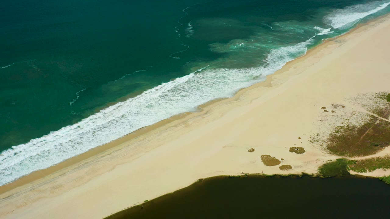 Overhead San Jose del Cabo beach view with the foam band spread across the middle of the frame, pale sand below it, dark inlet water at bottom, and no visible people.
