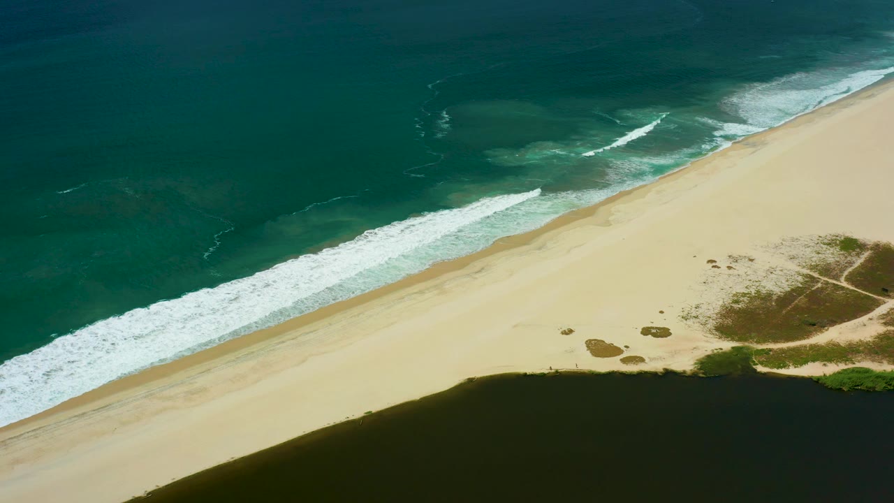 Aerial frame over San Jose del Cabo coastline with deeper teal water above, pale sand through the center, scattered vegetation to the right, and no visible people.
