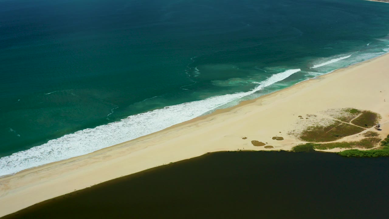 Aerial view of surf breaking along a sandy beach beside dark foreground water.