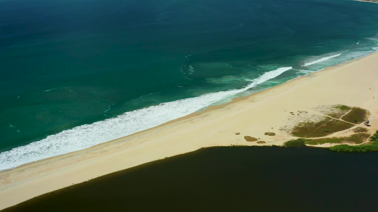San Jose del Cabo aerial frame with deeper blue water offshore, a white wave band along the sand, low vegetation near the beach edge, and no visible people.