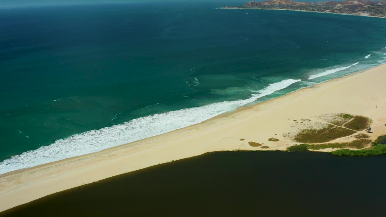 Mid-clip San Jose del Cabo aerial frame showing open ocean above a bright sand strip, breaking surf, a dark water band in the foreground, and no visible people.