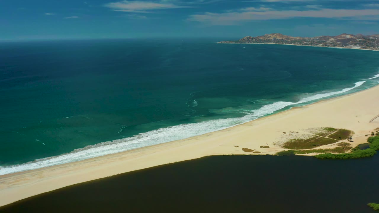 At 0:01, the San Jose Del Cabo coastline fills the frame with a diagonal sandy shore, rolling white waves, deep blue water offshore, distant hills, and no visible people.