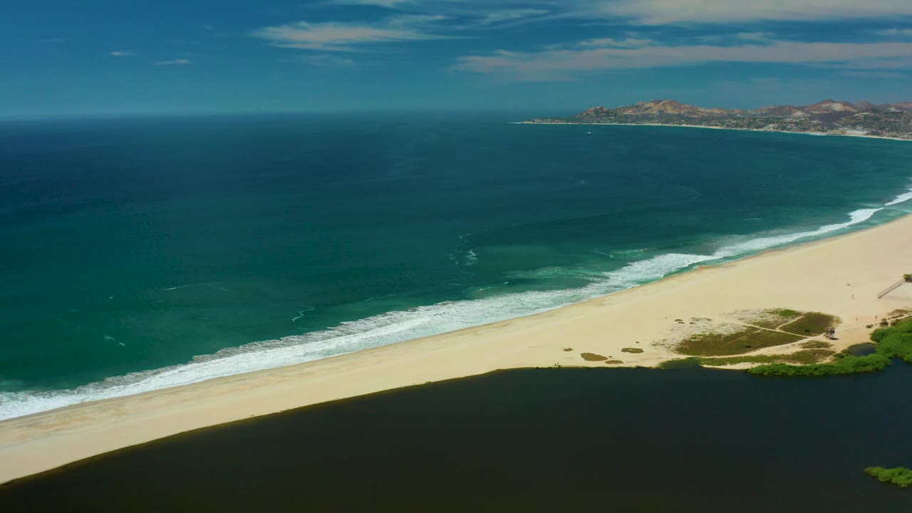 At 0:03, the San Jose Del Cabo aerial view holds on the shoreline as small wave lines approach the beach, with low hills and scattered clouds in the distance and no visible people.