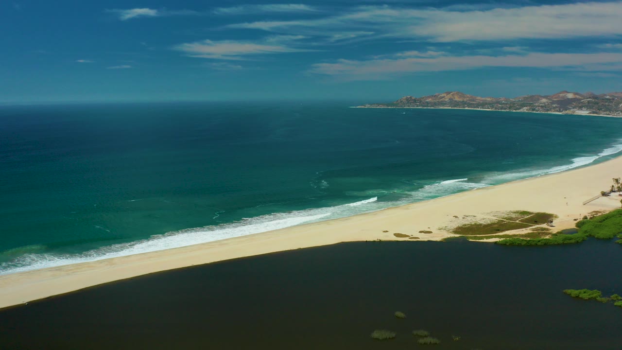 Aerial view in San Jose Del Cabo, Mexico, showing a sandy beach between turquoise ocean and a dark lagoon, with distant hills under streaked clouds and no visible people.