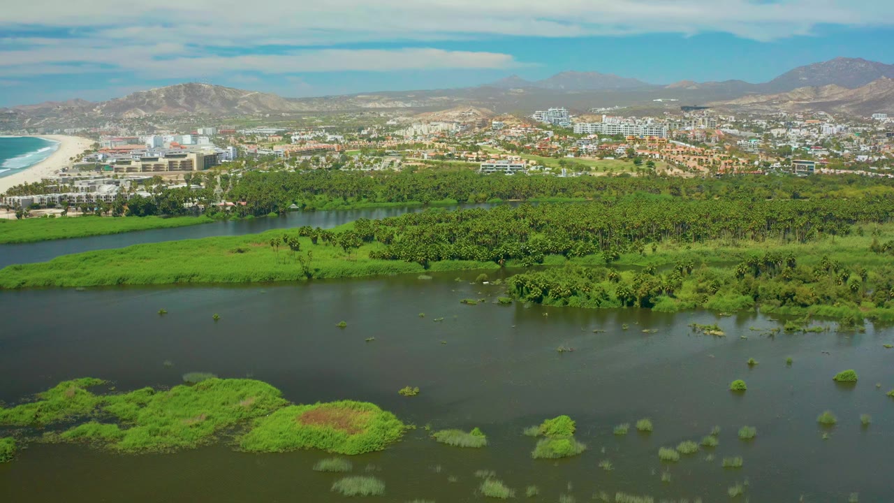 Wide aerial frame over San Jose del Cabo with dark river water and green marsh islands in front, a band of vegetation, beachside buildings, mountains, partly cloudy sky, and no visible people.