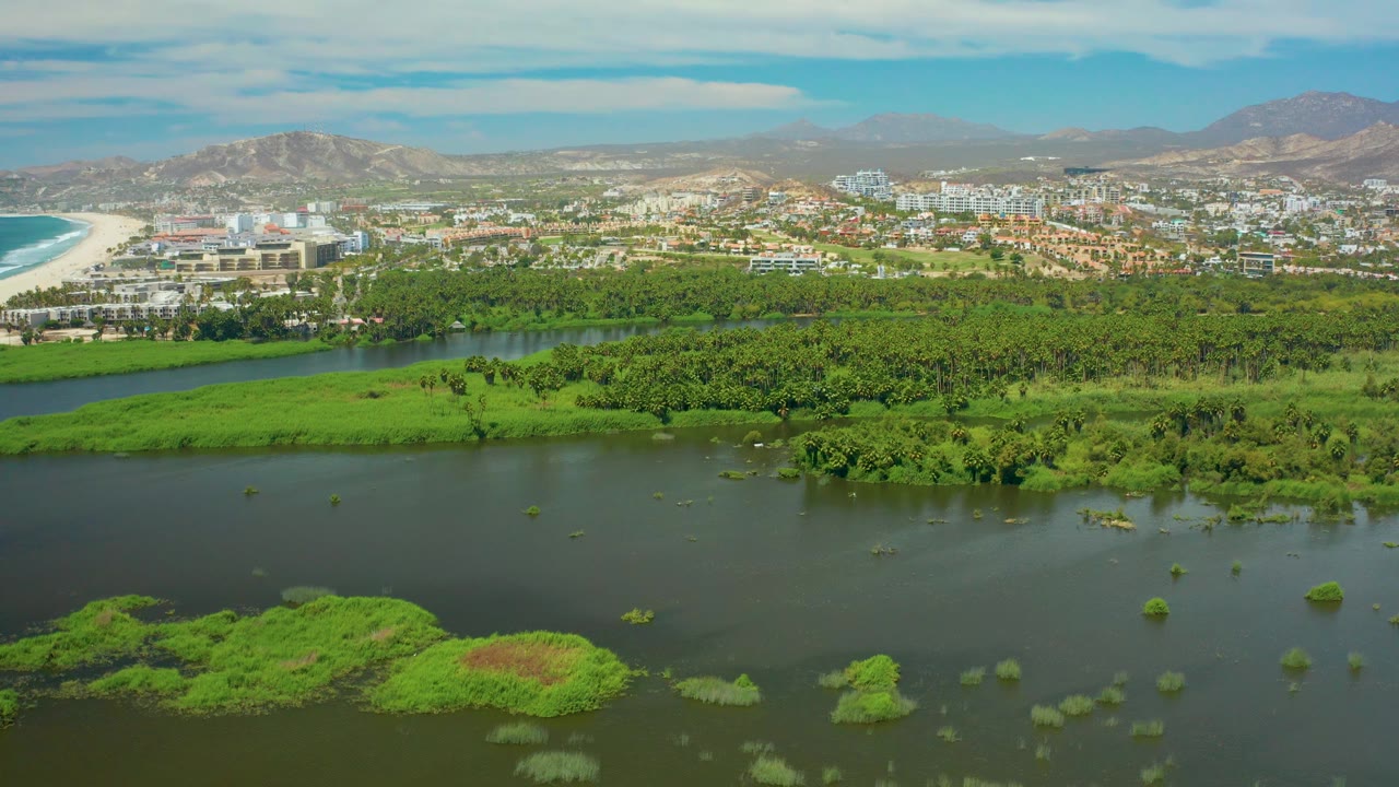 Aerial composition of marshy river foreground, green tree cover, beach buildings, and hills.